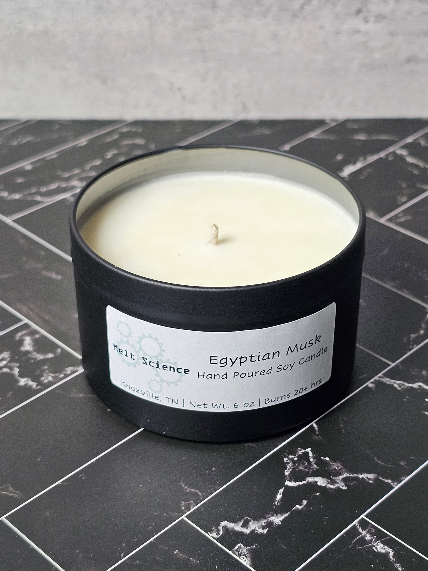 Candle in a black container with a label stating Egyptian Musk Hand Poured Soy Candle on a dark marble surface