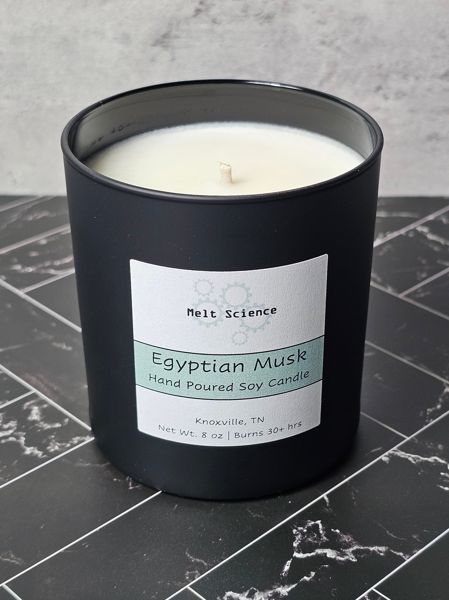 Black candle with a label stating Egyptian Musk Hand Poured Soy Candle on a gray and black marble surface